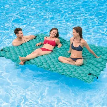 Three people in swimsuits—two women and one man—relax and smile while sitting on a large teal inflatable float mat in the water. The mat has a quilted texture and appears spacious enough to comfortably accommodate all three individuals in the pool