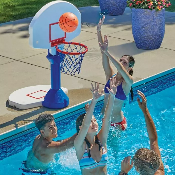 Four people are playing basketball in a swimming pool, with a blue and white portable hoop on the pool deck.