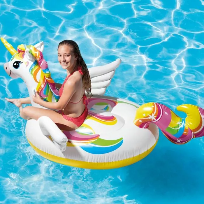 A smiling woman in a red bikini sits on a large white inflatable unicorn pool float with a rainbow mane and tail in a blue swimming pool.